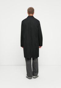 Hombre de pie frente a una pared blanca, vestido con un abrigo largo negro, pantalones grises y zapatillas blancas con rayas negras.