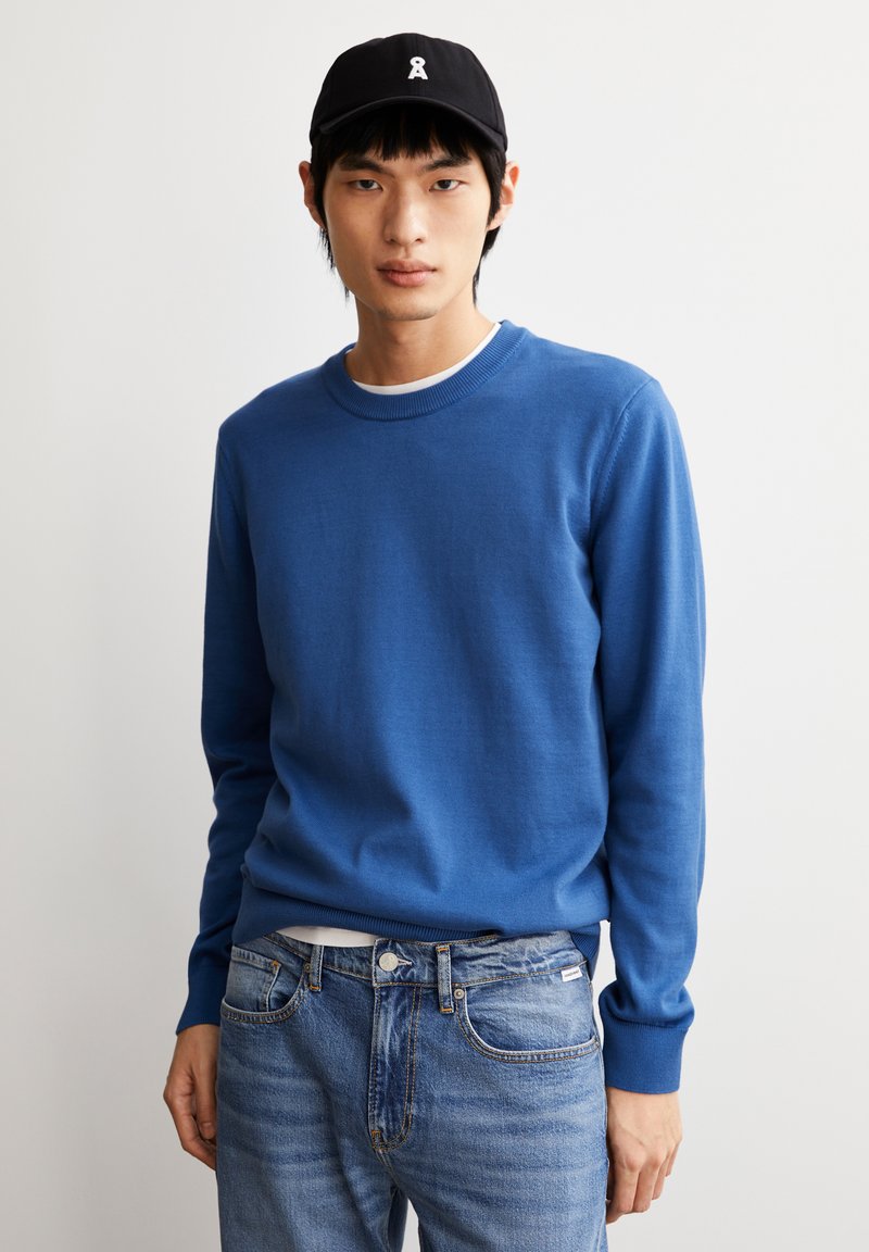 Blauer Baumwollpullover mit rundem Halsausschnitt und gerippten Bündchen, kombiniert mit hellblauen Jeans. Einfaches Design, keine Muster oder Akzente.