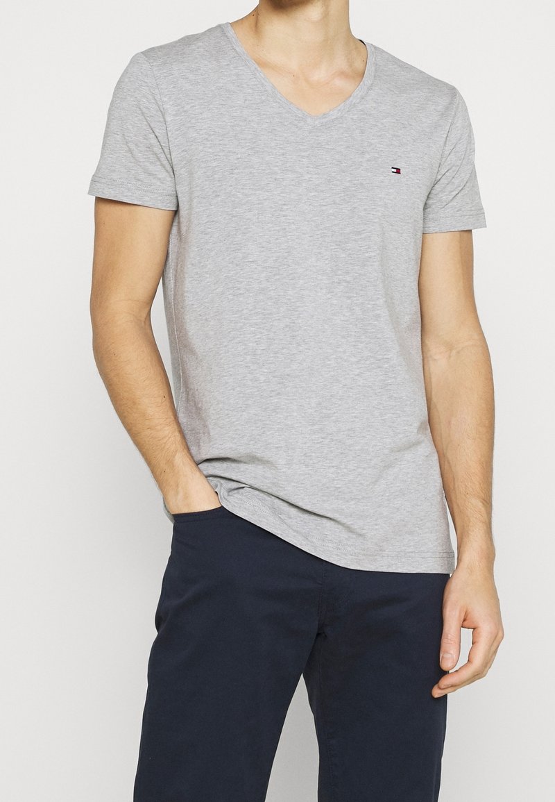 Graues Baumwoll-V-Ausschnitt-T-Shirt mit kurzen Ärmeln, mit einem kleinen Logo auf der Brust, kombiniert mit dunklen maritimen Hosen.