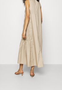 Robe maxi sans manches beige avec texture froncée à la taille, silhouette droite et tissu doux. Portée avec des sandales à talons de couleur claire.