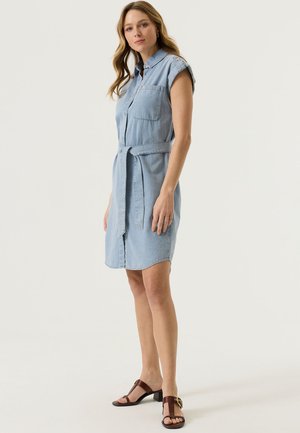 Femme portant une robe en denim sans manches bleu clair avec une ceinture, debout avec une jambe légèrement avancée, associée à des sandales à talons marron.