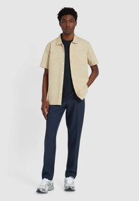 Lichtbeige overhemd met korte mouwen en een enkele borstzak, gecombineerd met marineblauwe broek en grijze sneakers. Glad stofoppervlak.