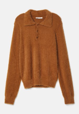 Person iført en fluffy brun langærmet polo, hvide vidde bukser og sorte chunky støvler, stående op ad en ensfarvet grå væg.