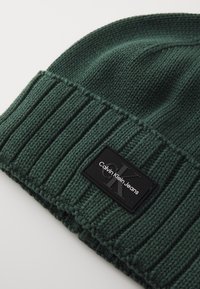 Beanie verde tricotat cu un design ribbed și un etichetă rectangulară neagră cu inscripția "Calvin Klein Jeans" în text alb.