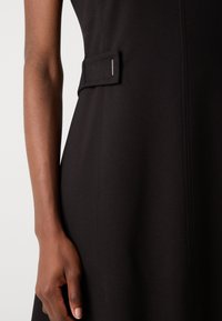 Black fabric dress featuring a structured silhouette, side pocket with a loop, and contrasting stitching. Smooth texture and minimalist design.
