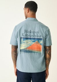 Svijetloplava košulja s kratkim rukavima od teksturiranog materijala na kojoj je na leđima istaknuta grafika planine Fuji i natpis "Hokusai Katsushika".