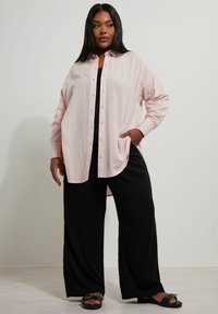 Ljust rosa knäppt skjorta med långa ärmar och sidfickor, bärs över en svart topp och vida svarta byxor, med platta sandaler.