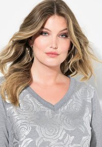 Top grigio a scollo a V con motivo floreale argentato, texture liscia e maniche corte; il modello ha i capelli mossi e indossa grandi orecchini a cerchio.