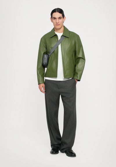 Chaqueta de cuero verde con diseño de cuello, combinada con pantalones gris con pliegues. El modelo también lleva un pequeño bolso cruzado negro.