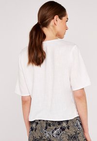 Weißes Leinen-T-Shirt mit kurzen Ärmeln, geradem Schnitt und rundem Halsauschnitt, kombiniert mit einem Rock im floralen Muster mit dunklen Tönen und hellen Akzenten.