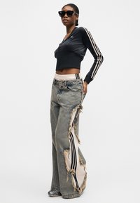 Γυναίκα με μαύρο cropped μπουφάν με φερμουάρ και λευκές ρίγες, και φθαρμένα φαρδιά τζιν που δείχνουν ριγωτό ύφασμα από κάτω.