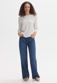Blouse blanche à manches longues avec un col arrondi, associée à un jean bleu évasé. Le modèle se tient les mains dans les poches, portant des chaussures beiges.