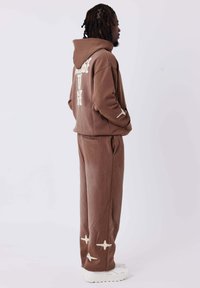 Felpa con cappuccio marrone con testo bianco sulla schiena, abbinata a pantaloni coordinati con accenti bianchi. Tessuto morbido con vestibilità rilassata.