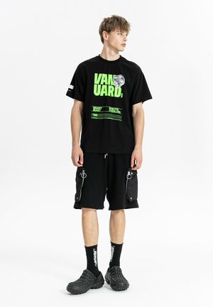 Joven con camiseta negra con diseño gráfico, pantalones cortos cargo negros con bolsillos con cremallera, calcetines negros y zapatos negros gruesos, de pie sobre fondo blanco.