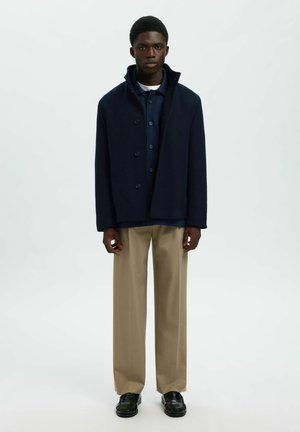 Jaunas vyras stovi vilkėdamas tamsiai mėlyną švarką ant mėlynos marškinių, smėlio spalvos kelnių ir juodų loafers batų fono prieš paprastą baltą sieną.