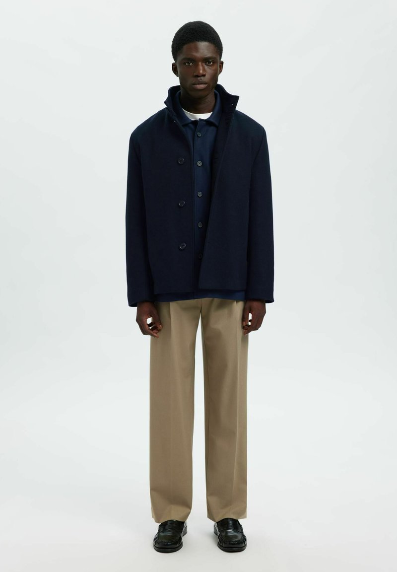 Mladi muškarac stoji u mornarsko plavoj jakni preko plave košulje, bež hlačama i crnim mokasinkama, naslonjen na jednostavnu bijelu pozadinu.