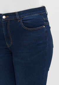Dark blue denim jeans featuring a mid-rise waist, five-pocket design, and contrasting orange stitching details. Smooth texture with minimal distressing.