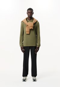 Polo verde oliva a maniche lunghe con un maglione beige annodato intorno al collo, abbinato a pantaloni neri larghi e sneakers con suola stampata.