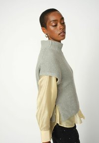 Lichtgrijze gebreide trui met een hoge kraag en korte mouwen, gelaagd over een lichtgele blouse met lange mouwen. Textuurpatroon details zijn zichtbaar.