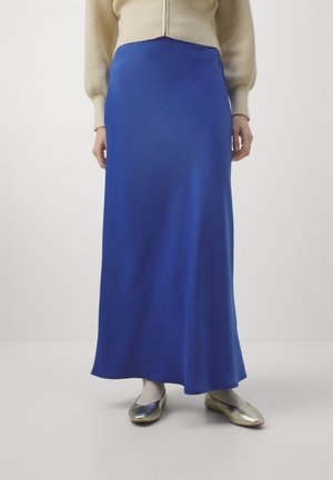 Donna con gonna lunga blu, maglione beige, collant chiari e scarpe basse argentate lucide, in piedi su sfondo uniforme.