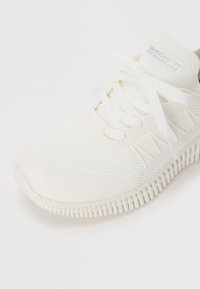 Baskets en mesh blanches avec un bout rond, présentant un design texturé, des lacets blancs et une semelle froncée. Détail du logo sur la languette.