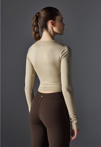 Langermet tettsittende topp i beige med en glatt tekstur og rund hals, kombinert med høyhlsede brune leggings med subtil logo-detalj.