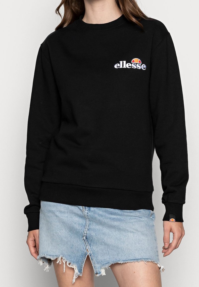 Sudadera negra con cuello y puños acanalados. Cuenta con un logo bordado de "ellesse" y un acento colorido. Combinada con una falda de mezclilla desgastada.