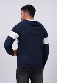 Hoodie marine avec des rayures blanches sur les manches, en matériau coton doux, poches kangourou, capuche réglable et poignets côtelés.