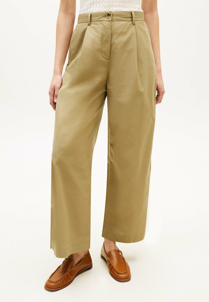 Persona con pantaloni beige a gamba larga e mocassini in pelle marrone, in piedi con le mani rilassate ai lati contro uno sfondo neutro.