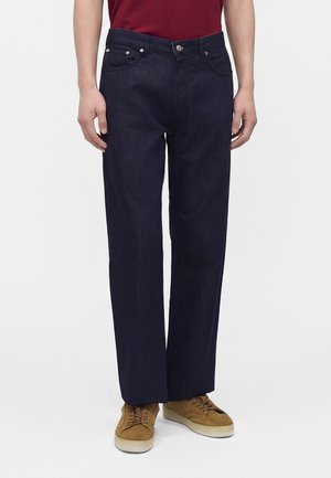 Person iført mørkeblå jeans med lige ben, brune ruskindssneakers og en rød skjorte, stående mod en ensfarvet hvid baggrund.