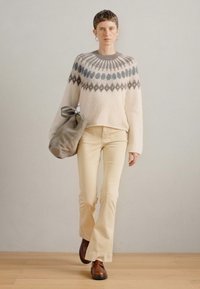 Κροκετέ πουλόβερ σε κρέμα με μοτίβο στο γιακά, μπεζ φαρδιά παντελόνια και καφέ παπούτσια. Κρατώντας μια γκρι τσάντα, στέκεται μπροστά σε ουδέτερο φόντο.