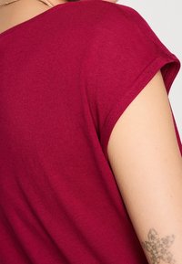 Femme portant un t-shirt bordeaux à manches courtes affichant un tatouage à motifs floraux sur le haut de son bras.