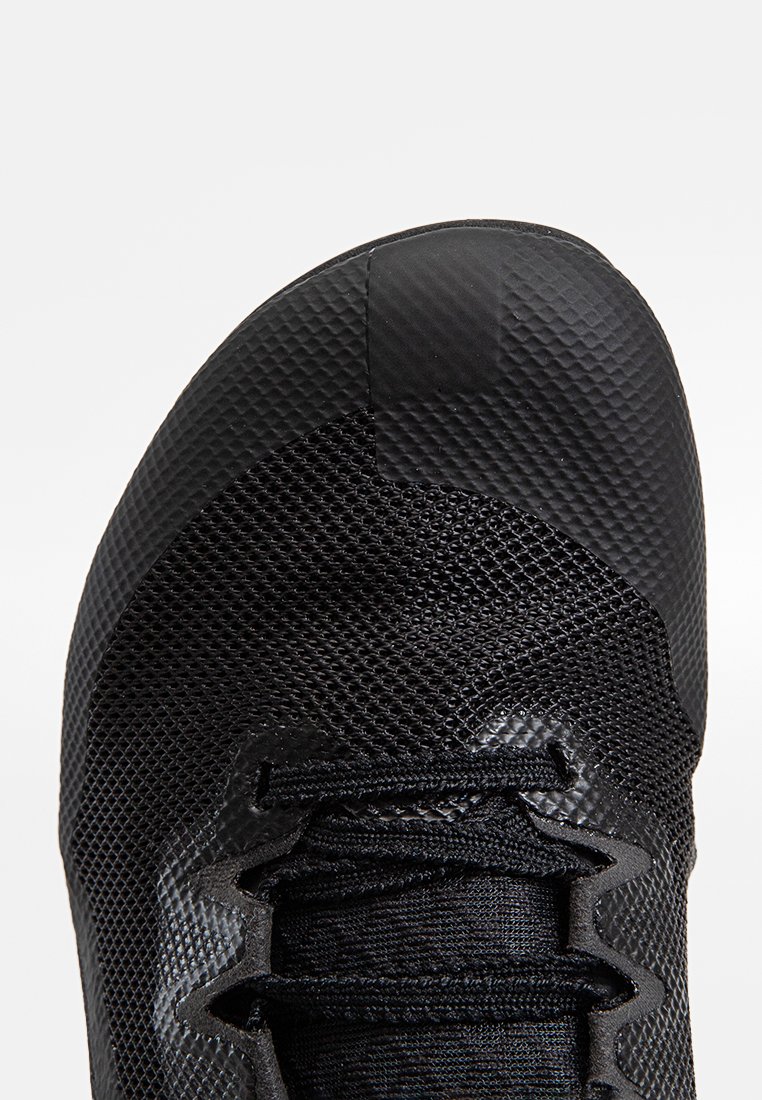 Zbliżenie czarnego obuwia sportowego z widoczną siateczką, teksturowaną osłoną oraz sznurówkami na jednolitym tle.