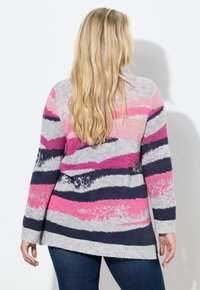 Donna bionda che indossa un maglione a maniche lunghe a righe rosa, grigio e blu navy con jeans, vista da dietro contro un muro bianco.