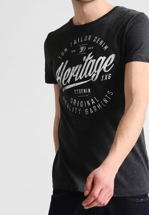 Mężczyzna w ciemnoszarej koszulce z białym, stylizowanym na vintage napisem "Heritage" oraz dodatkowymi mniejszymi napisami "Tom Tailor Denim" i "Original Quality Garments".