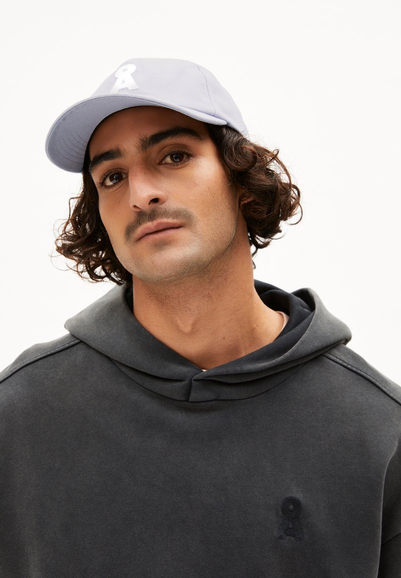 Lichtpaarse baseballcap met een wit geborduurd logo, gedragen met een donkergroene hoodie met een subtiele logodetail.