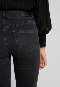 Jeans de denim negro de cintura alta, con dos bolsillos traseros y una textura suave. Las costuras visibles añaden detalle al diseño.