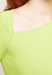 Haut à manches longues vert clair côtelé avec un large décolleté et un tissu texturé, présentant une silhouette ajustée et des détails de coutures subtiles.