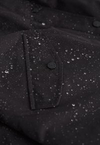 Czarny wodoodporny materiał z kroplami wody na powierzchni. Posiada kieszeń z czarnym guzikiem. Gładka tekstura z minimalnym połyskiem.