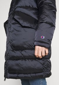 Chaqueta acolchada negra con una textura brillante y suave, que presenta un gran bolsillo en el lateral y un pequeño logo bordado en el puño.