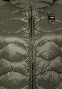 Pufferjack in donkergroen olijf, met een golvende doorgestikte patroon, een voorrits en een label op de borst waarop de merknaam duidelijk zichtbaar is.