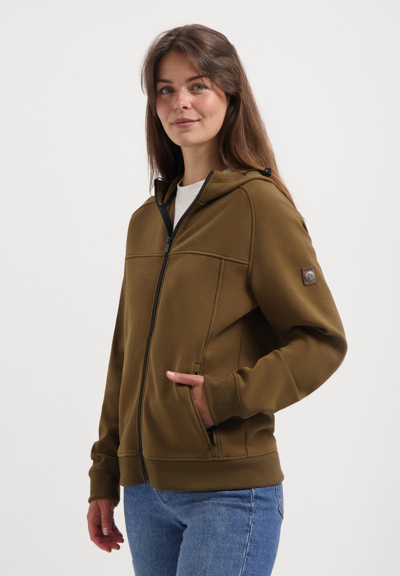 Jeune femme portant un sweat à capuche zippé marron avec les mains dans les poches, associée à un jean bleu, se tenant contre un fond blanc uni.