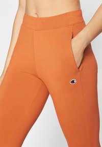 Leggings laranja elásticos com cintura alta, apresentando bolsos laterais e um pequeno destaque de logotipo no quadril. Teixeira de tecido suave e lisa.
