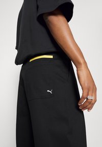 Pantaloni neri con una tasca posteriore, dotati di un logo Puma bianco e un accento giallo in vita. Tessuto morbido con un design a taglio dritto.