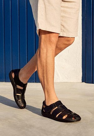 Mann trägt beige Shorts und schwarze Ledersandalen und geht auf hellem Gehweg vor einem blau-weißen Wandhintergrund.