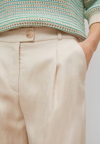 Beżowe, gładkie spodnie z materiału z plisowanym przodem i bocznymi kieszeniami. Wyposażone w jeden okrągły guzik w pasie. Powyżej sweter z teksturowanego dzianiny.