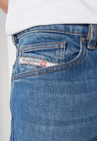 Primer plano de un bolsillo de jeans de mezclilla azul con costuras naranjas y etiqueta de la marca Diesel Industry en blanco y rojo.