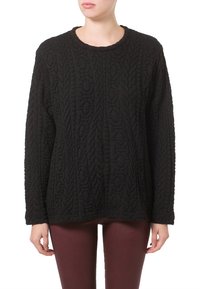 Mujer vestida con un suéter negro de punto trenzado y de manga larga, y pantalones rojo oscuro, de pie contra un fondo blanco.