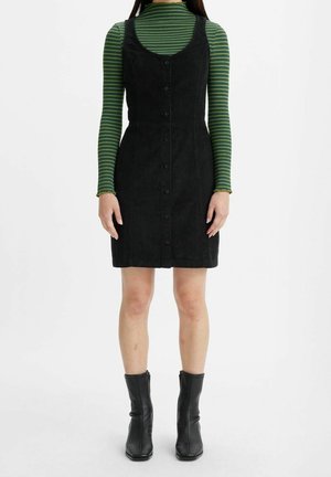 Person trägt ein schwarzes, ärmelloses Kleid mit Knöpfen über einem grün gestreiften Langarmshirt und schwarzen Stiefeletten, steht vor einem weißen Hintergrund.