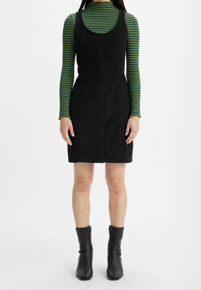 Personne portant une robe noire sans manches boutonnée par-dessus un haut à manches longues rayé vert, avec des bottines noires, debout sur un fond blanc.